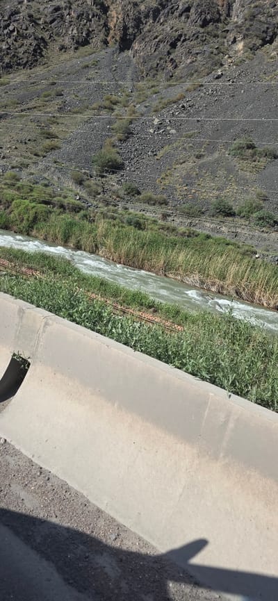 The image depicts a rugged, natural landscape featuring a fast-flowing river bordered by dense green vegetation. In the foreground, a concrete barrier runs along the edge of the scene, with a gravelly surface and shadows cast by an unseen object. The river, with its white, churning water, cuts through the frame, flanked by tall reeds and other green plants. Beyond the riverbank, a steep, rocky hillside dominates the background, characterized by dark, loose stones and sparse vegetation. Several thin, dark lines, possibly power or utility lines, are visible stretched across the hillside. The overall environment suggests a remote or undeveloped area, possibly in a mountainous or semi-arid region, consistent with the general geography of Kyrgyzstan.
