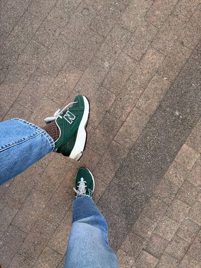 A forensic overhead photograph captures the lower extremities of an individual standing on a paved surface in Saint-Jean-de-Luz, France. The subject wears light-wash blue denim jeans and brown ribbed socks, paired with dark green New Balance sneakers. The sneakers feature white 'N' logos, white laces, and white midsoles, with black outsoles partially visible. The left foot is positioned forward and slightly raised, displaying the shoe's lateral profile, while the right foot is planted, revealing its dorsal aspect. The ground is composed of rectangular, reddish-brown concrete pavers with an exposed aggregate finish, arranged in a diagonal pattern. A distinct linear shadow extends diagonally across the pavement from the upper right to the lower left, indicating a direct light source.