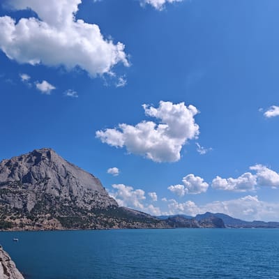 The image depicts a coastal landscape with a prominent rocky mountain range on the left, bordering a body of turquoise water. The mountain is rugged and covered with sparse vegetation. The water is calm, with small ripples. In the distance, the coastline stretches out with more mountains and some scattered buildings. The sky is a vibrant blue, dotted with fluffy white clouds. A small white boat is visible on the water to the left of the mountain. The scene appears to be a scenic view of the Black Sea coast, possibly near Sudak, Ukraine, given the characteristic mountainous terrain and coastline often seen in that region.