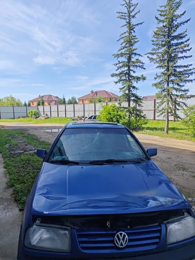 A blue Volkswagen car with a cracked windshield and significant front-end damage is parked outdoors on a dirt and grass area. The car's hood is dented and crumpled, and one headlight is missing. In the background, there is a metal fence, several houses with red roofs, and two tall evergreen trees. The sky is blue with scattered clouds. The scene appears to be located in a suburban or rural area of Оқжетпес, Kazakhstan.