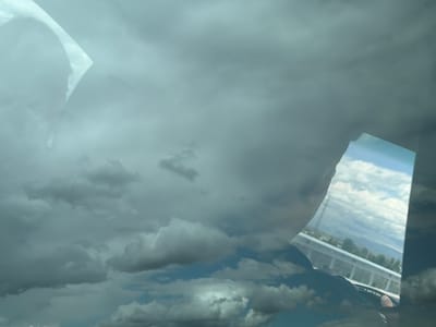 The image shows a cloudy sky with reflections of a bridge and landscape on a car window. The bridge railing is white, and hints of trees and a distant mountain are visible in the reflections. A person's head, wearing a hat, can be seen reflected on the window. The rest of the image is filled with various cloud formations against a grey-blue sky. There is no definitive information present to confirm that the image was taken in Agarak, Armenia.