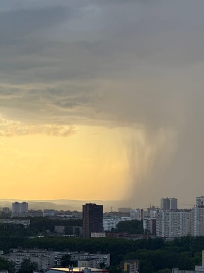 The image depicts an urban landscape in Shirkovo, Russia, under a dramatic sky. The foreground is dominated by residential buildings and dense green trees. In the mid-ground, more buildings of varying heights are visible, including a prominent tall, dark-colored apartment building. The background shows a hazy cityscape with more distant buildings and rolling hills. A striking feature is a heavy rain shower occurring in the distance, illuminated by the warm, golden light of the setting sun on the left side of the frame. The sky above is a gradient of gray and yellow, with dark clouds on the left transitioning to a lighter, overcast sky on the right. No people are clearly visible in the image. The overall atmosphere is one of an impending or ongoing storm over a city.