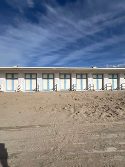 The image shows a row of beach huts in Trouville-sur-Mer, France. The huts are numbered and have blue and white doors. They are located on a sandy beach, with the ocean and sky visible in the background. The sky is blue with white clouds. The beach is made of light brown sand and there are footprints visible in the sand.