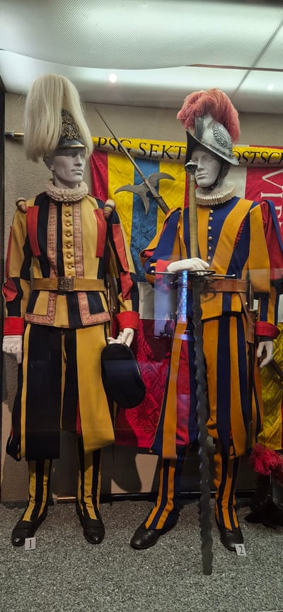 This media file displays two mannequins dressed in the traditional uniform of the Swiss Guard, likely part of a museum exhibit. The uniforms are highly decorative, featuring alternating stripes of bright yellow and dark blue on trousers and shirts, with red accents on cuffs and collars. The upper mannequin wears a metallic helmet adorned with a large, pink feather plume. It holds a long, ornate halberd. The lower mannequin sports a black cap with a red band and a stylized blonde wig. Both mannequins are posed standing, with their weapons held in front of them.
Behind the mannequins, a large banner with bold red and yellow stripes is visible. Text on a vertical yellow band reads "PSC SEKT". A small white tag with the number "2" is affixed to the right leg of the upper mannequin, and a similar tag with "1" is visible on the lower mannequin's left leg. The mannequins are displayed within a glass case, suggested by reflections on the left side of the image. The background is a textured, grey surface. The scene appears to be indoors, possibly a historical exhibit in Sargans, Switzerland, given the context provided. The lighting is artificial, casting shadows and highlighting the vibrant colors of the uniforms. The overall impression is one of historical display and military regalia.
