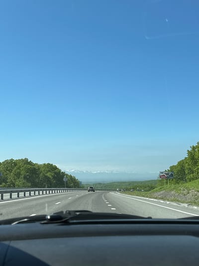 An eye-level, forward-facing shot captures a car's dashboard and windshield view of a highway on a clear, sunny day. The highway, with multiple lanes and white dashed lines, curves to the right, bordered by a metal guardrail and lush green trees on either side. In the distance, snow-capped mountains rise majestically under a vast, bright blue sky. Several cars are visible on the highway, moving in the same direction. A billboard with text and images stands on the right side of the road. The scene suggests a road trip through a scenic, mountainous region, possibly during spring or summer. The location is identified as the city of Svetly, Russia.