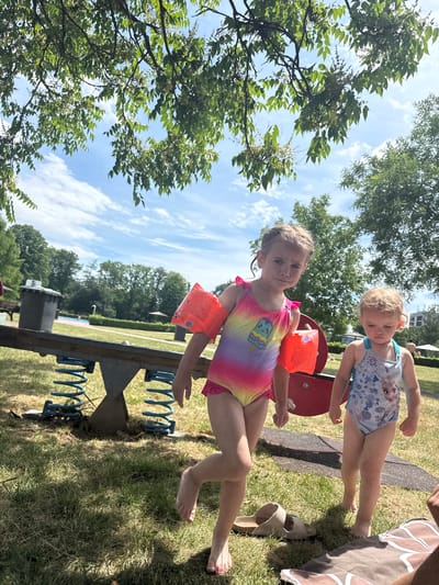 This outdoor photograph, taken in Giessen, Germany, captures a sunny day with a bright blue sky and scattered white clouds. In the foreground, two young children are the main subjects. The child on the left, wearing a colorful rainbow swimsuit with an ice cream print and orange arm floaties, is looking towards the camera with a slight smile. Her braided hair is visible. To her right, a younger child with blonde hair is wearing a light blue swimsuit with a cartoon character, possibly from Frozen. This child is leaning forward, looking down.

They appear to be playing on a red and metal playground structure, possibly a seesaw or a spring rider, which is partially visible in the lower portion of the image. The ground is covered in green grass, with some dry patches. In the background, trees and more playground equipment can be seen, suggesting a park or a recreational area. A pair of beige sandals are visible on the grass near the right edge of the frame. The image has a slightly tilted perspective, adding a dynamic feel to the scene. The overall mood is cheerful and indicative of a summer day spent outdoors. No legible text is visible on the children's clothing or arm floaties beyond small print.