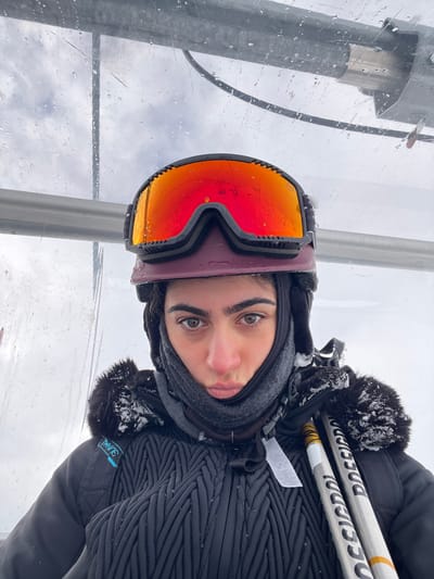 A person wears ski goggles and a balaclava, positioned slightly askew, with ski poles visible. They are in an enclosed space, possibly a gondola or ski lift, with water droplets on the transparent barrier. The person is wearing a dark, quilted jacket with fur trim. The background shows a cloudy, snowy scene.