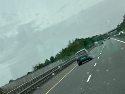 An oblique, slightly tilted, medium shot captures a view from inside a vehicle on a highway under a cloudy sky. The scene is likely on the M11 motorway near Gorey, Ireland, given the visible road markings, barrier, and surrounding greenery, consistent with Irish highway infrastructure. In the foreground, the grey asphalt road features a single yellow line to the left and intermittent white dashed lines indicating lanes. Several water droplets or smudges are visible on the surface the photo was taken from, obscuring parts of the scene.
A dark-colored hatchback vehicle, possibly a Peugeot, is the primary subject, traveling away from the camera in the right lane. Its red taillights are illuminated. Further down the highway, other vehicles are present but less distinct due to distance and motion blur.
To the left, a concrete barrier with a metal guardrail on top separates the highway from the adjacent embankment. This embankment is covered with lush green trees and foliage, suggesting a rural or semi-rural setting. The sky is overcast, with varying shades of grey and some lighter patches, indicating a dull day, possibly with light rain. The overall impression is one of routine travel on a highway in Ireland.