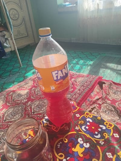 A Fanta bottle and a glass jar sit on a red patterned blanket on a green carpet. The Fanta bottle is partially full of a red liquid and has a yellow cap. The glass jar is empty and to the left of the bottle. In the background, a door with ornate carvings is visible. Sunlight streams in from a window covered by sheer curtains. The scene appears to be indoors, possibly in a home in Kasansay, Uzbekistan, given the patterned textiles and carpet.