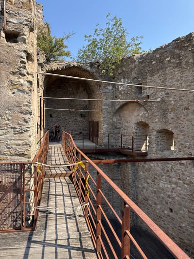 A sunlit scene shows a wooden walkway with weathered, rusty railings leading into the stone ruins of a castle structure, possibly located in Dolceacqua, Italy. The walkway, constructed of wooden planks, has a series of vertical supports casting long shadows across its surface. Two horizontal ropes are tied to the railings, one with yellow warning tape attached. In the background, a cutout figure of a person in historical costume stands near an arched entryway. The ancient stone walls of the castle are partially overgrown with green foliage, and the clear blue sky is visible above.