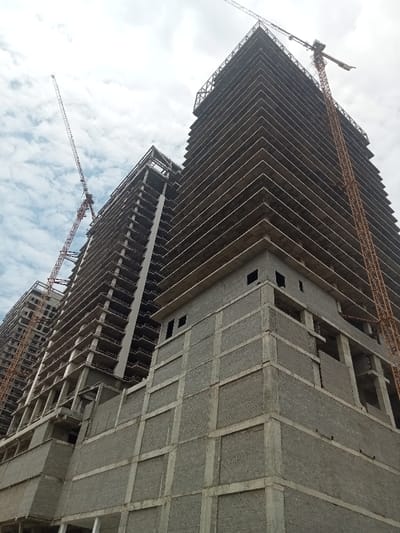 This image captures a wide-angle view of a construction site featuring two towering, unfinished high-rise buildings under a cloudy sky in Luanda, Angola. The perspective is from below, looking up, emphasizing the immense scale of the structures.
The primary subjects are the two concrete skeletal frames of future skyscrapers. The building on the right is more prominent and appears taller, with numerous concrete floor slabs already cast, creating a repeating pattern of horizontal lines across its facade. A large yellow-orange tower crane stands next to this building, extending well above its current height, indicating ongoing construction.
To the left, a second, slightly less advanced high-rise is visible, also with its concrete framework in place. Another slender red and white tower crane is positioned near this building, reaching high into the sky.
In the foreground, part of one of the buildings (likely the one on the right or a connecting structure) features completed sections of external walls made of light grey, block-like material, possibly concrete blocks or precast panels, forming a grid pattern. These sections have small, dark, rectangular openings, likely for windows or vents. Above these finished sections, the building reverts to its bare concrete frame with exposed floor slabs.
The sky is overcast with diffused light, suggesting a cloudy day, but it remains bright. There are no people visible, reinforcing the focus on the large-scale construction itself. The scene conveys a sense of significant urban development and growth. No visible text is present in the image.