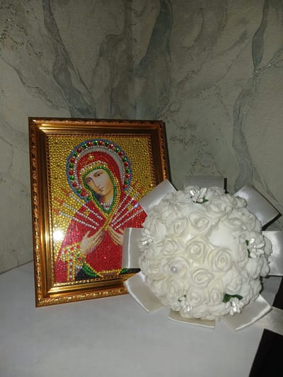 A framed diamond painting of the Virgin Mary rests on a white surface next to a white rose bouquet. The painting, with a gold frame, features a depiction of the Virgin Mary adorned in red and gold, with a halo of colorful gemstones. The bouquet, composed of numerous small white roses and accented with white ribbons and small sparkling details, is positioned to the right of the painting. The background is a textured wall with a marble-like pattern in shades of gray and blue. The lighting suggests an indoor setting, likely a home environment, and the overall scene appears to be a still life arrangement. There is no indication of the specific location within Prokudskoye, Russia.