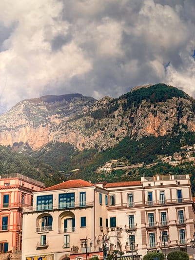 The image displays a scenic view of a mountainous coastal town. In the foreground, several multi-story buildings with white and salmon-pink facades line a waterfront area. These buildings feature arched windows, balconies with railings, and red-tiled roofs. The architecture suggests a European style, likely Mediterranean. Behind the buildings, a steep, rocky mountain covered in dense green vegetation rises dramatically. The mountain is characterized by sheer cliffs and a rugged, textured surface. The sky above is filled with dramatic, cloudy formations. There are no visible people or specific actions taking place in the scene. The lighting indicates it is daytime, with the sun casting shadows and illuminating the mountain face. The overall impression is one of natural beauty combined with a charming, built environment. There is no evidence to suggest this location is in Silvolde, Netherlands.