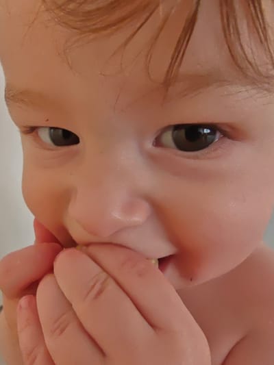 The image is a close-up shot of a young child's face. The child has light-colored hair and is looking towards the viewer with a slight smile. The child's hands are near their mouth, and it appears they might be eating something. The background is out of focus and appears to be a plain, light-colored wall, suggesting an indoor setting. There is no visible information within the image to indicate a location in Georgsmarienhütte, Germany.
