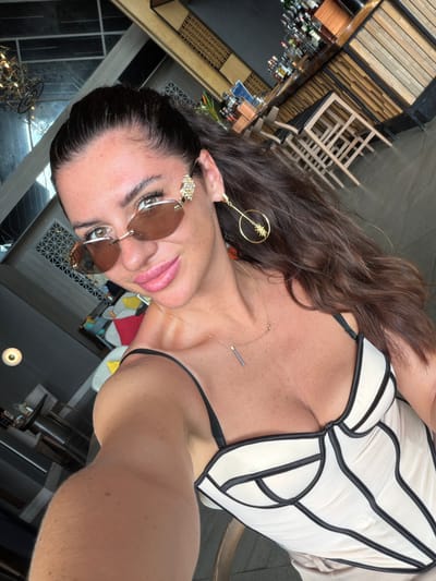 A woman is captured in a selfie, showcasing a close-up view of her upper body and face. She is wearing a light-colored corset-style top with black trim, and a delicate gold necklace with a small bar pendant. Her hair is long and dark brown, falling over her shoulder. She wears oversized, round, brown-tinted sunglasses with a jeweled embellishment on the side, and a dangling earring with a starburst design and a jeweled accent. Her makeup is visible, with defined eyebrows, eyeliner, and lipstick.
The background appears to be an indoor setting, possibly a restaurant or bar, with wooden shelves holding bottles visible to the left. The lighting suggests daytime. The woman's expression is a slight smile, with her head tilted. The image is oriented sideways.