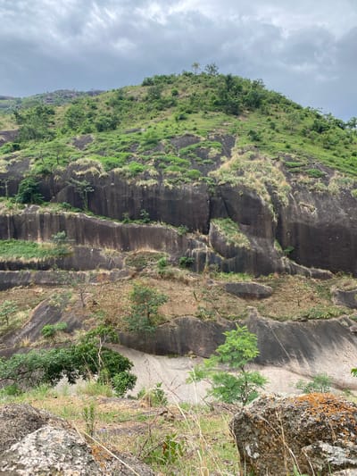 The image shows a steep, rocky hillside in Ganawuri, Nigeria. The hillside is covered in patches of green vegetation, including grass, shrubs, and trees. The rock face is dark grey and has several vertical grooves or ridges. The sky is overcast. A large boulder covered with lichen and dry grass is visible on the right side of the image.