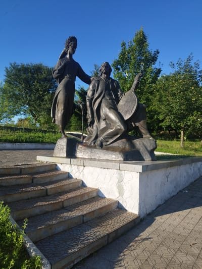The image shows a bronze monument of two figures in Novyi Rozdil, Ukraine. The monument is set on a tiered base of stone and concrete, with steps leading up to it. The figures are positioned on a rough, rocky base. To the left stands a female figure, draped in a long gown, with her left hand resting on the shoulder of the male figure. She appears to have a wing or plume on her head. To the right, a male figure is seated, wearing a long coat and holding a stringed instrument, possibly a kobza or bandura. He has a beard and a thoughtful expression. Behind the monument, a blue sky dominates, with green trees forming a backdrop. The ground in front of the monument is paved with patterned tiles, and a section of green foliage is visible to the left of the steps. A lamppost stands behind the male figure.