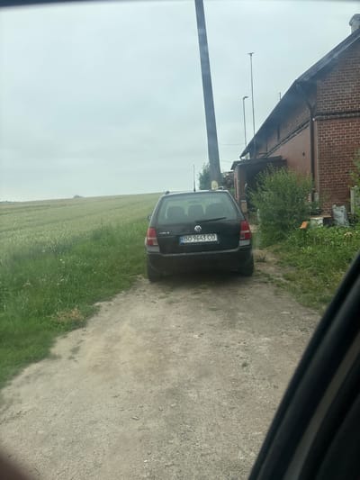A black Volkswagen Golf estate car is parked on a gravel path in front of a brick building with a dark roof. The car's license plate reads "BO 9643 CO". To the left of the car is a green field. The sky is overcast. The building appears to be a residential or agricultural structure. In the background, there are trees and telephone poles.