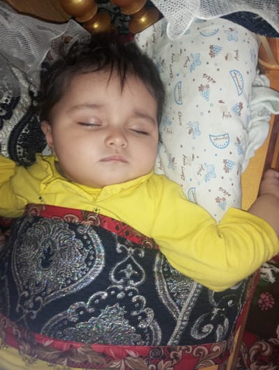 A sleeping infant is depicted in a close-up, top-down view, positioned indoors, presumably in Galaosiyo, Uzbekistan. The infant, wearing a yellow long-sleeved top, lies on its back with eyes closed. Its head rests on a white pillow adorned with blue and brown stylized fruit (apples, cherries, grapes, watermelons) and bear figures. A dark blue or black blanket with shimmering silver, possibly holographic, intricate paisley and floral patterns covers the infant's lower body. A red and brown patterned fabric is visible beneath the blanket and along the edges of the frame. To the upper left, a translucent white mesh fabric and a golden, curved decorative object are partially visible. The infant's facial expression is neutral, indicative of sleep.