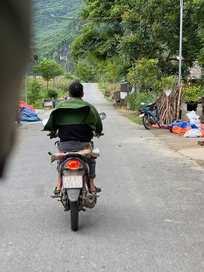 In Lùng Tám Commune, Vietnam, a person is riding a motorbike on a paved road. The rider is wearing a green jacket over a black shirt and grey pants. The motorbike has a blue license plate with white text that reads "23-B1 176.82". The road is lined with trees, foliage, and some structures on the side. In the background, a steep, lush green mountainside is visible. There are also stacks of wood, tarps, and bags on the side of the road, suggesting a rural or semi-rural setting. The scene is captured from a slightly elevated perspective, likely from another moving vehicle.