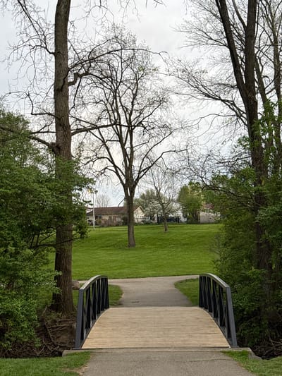 The image depicts a park scene, with a bridge spanning a green grassy area. The bridge has a wooden deck and dark metal railings. A paved pathway leads onto and off the bridge. Bare trees are visible on either side of the path, some with sparse new foliage. In the background, houses are seen across the lawn. The location is Grove City, Ohio, USA.