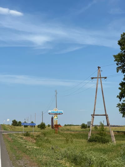 This is a vertical, slightly tilted photograph of a rural landscape under a clear blue sky with thin, wispy clouds. The foreground is dominated by tall green grass and vegetation. A paved road runs along the bottom right of the image, leading towards the horizon.
Several telephone poles with wires are visible, spaced along the roadside. One prominent pole in the middle distance supports a sign that reads "С. ЕНИСЕЙСКОЕ" (S. ENISEYSKOE) with a sun graphic and "ОСНОВАНО В 1777 ГОДУ" (FOUNDED IN 1777) below it. Further down the road, another blue sign points to the left, indicating "Б-ЕНИСЕЙСКОЕ 4" (B-ENISEYSKOE 4). In the distance, trees and more vegetation are seen, with a hazy horizon.
The overall impression is one of a sunny day in a rural area of Russia, likely during the daytime given the bright sunlight and clear skies. There are no people visible in the frame. The scene appears peaceful and somewhat deserted.