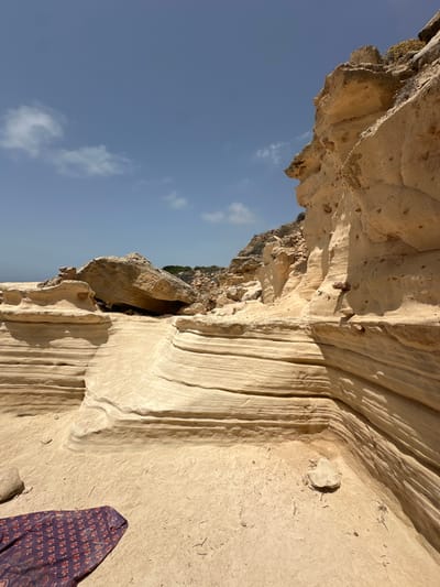 A bright, sunny day is depicted in a natural setting featuring unique rock formations along a coastline. The foreground is dominated by layered, sandy-colored rock that has been sculpted by erosion, creating horizontal striations and hollows. To the left of the foreground, a folded piece of fabric, appearing to be a patterned scarf or sarong in shades of deep purple, red, and white, lies on the sandy ground.

The midground consists of larger rock formations, including a significant boulder balanced precariously on smaller eroded rocks. Behind these, a rugged, rocky hillside rises, dotted with sparse vegetation. The background is a clear blue sky with a few wispy white clouds. The overall environment suggests a coastal area with distinct geological features, likely a beach or cove. The scene is located in Bellavista, Spain. There are no people or animals visible in the image.