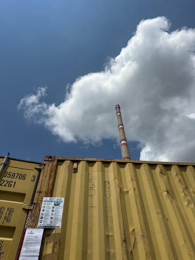 This media file presents an industrial landscape, rotated ninety degrees counter-clockwise. When viewed correctly, a tall, slender smokestack stands centrally against a bright blue sky, partially obscured by large, fluffy white clouds. The smokestack is cylindrical and features alternating beige/white and red bands.

In the foreground, two large, weathered yellow shipping containers occupy the right side of the frame. The upper container shows significant wear, with dents and scratches on its corrugated metal surface. The lower container is partially visible, also yellow and corrugated, with several labels and markings.

The sky is a clear, vibrant blue, with a dynamic arrangement of white cumulus clouds, suggesting a sunny day with good visibility. The overall setting implies a facility related to heavy industry or power generation, characteristic of areas like Pirdop, Bulgaria, known for its industrial sites.

Visible text includes:
On the lower shipping container: "359706", "22G1", "8 KGS", "O LBS", "KGS", "Los", "L", and "S".
A "SAFETY RULES" sign is affixed to the lower container, titled "SYNERGY ПРАВИЛА ЗА БЕЗОПАСНОСТ" (SYNERGY SAFETY RULES) and featuring various safety pictograms and warnings.
Another document next to it shows "SYNERGY", "УТВЪРЖДАВАМ" (Approved), and "ИНСТРУКЦИЯ №" (Instruction No.).
The upper shipping container has partially visible text that appears to include "MEDITE".
No people or active events are captured in the static scene.