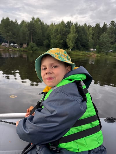 The image shows a young boy in a boat on a lake. He is wearing a yellow and blue patterned hat and a blue jacket over a bright green life vest. The boy is looking towards the camera with a slight smile. The water is dark and reflective, showing the trees on the shore and a cloudy sky. The trees form a dense line along the background, with some structures or buildings partially visible among them. The overall impression is of an outdoor recreational activity on a possibly overcast day. There is no visible text in the image.