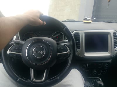 The image shows the interior of a Jeep vehicle, specifically the dashboard and steering wheel, as viewed from the driver's perspective. A person's hand is visible holding the steering wheel. The steering wheel has the Jeep logo in the center and various buttons for controlling audio and cruise control. The dashboard features a central infotainment screen, air vents, climate controls, and a gear shifter. A small, silver toy helicopter is perched on the dashboard. The scene appears to be set outdoors, with a light-colored wall visible through the windshield. The vehicle is likely parked in Yeghvard, Armenia, based on the provided context, though this cannot be definitively confirmed from the image alone.