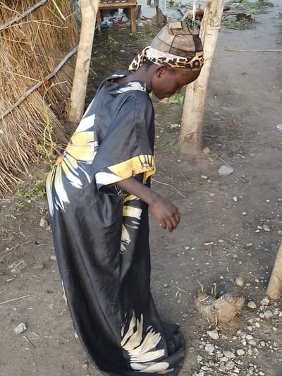A young Black person is standing outdoors, facing left, and looking down. They are wearing a long, black, flowing garment with a yellow and white floral pattern. On their head, they have a cap made of patchwork fabric and a leopard-print headscarf. The person's right hand is visible, resting slightly away from their body. The ground beneath them is dirt with scattered rocks and some dry vegetation. In the background, there are several wooden posts and a structure constructed from dried grass. The environment appears to be rustic and rural.