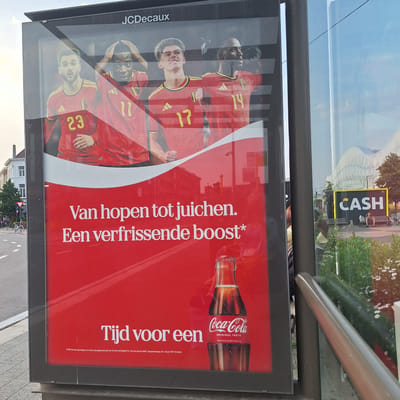 The image shows a Coca-Cola advertisement at a bus stop in Mechelen, Belgium. The advertisement features four soccer players in red jerseys with yellow numbers. The text on the advertisement reads "Van hopen tot juichen. Een verfrissende boost." and "Tijd voor een". A bottle of Coca-Cola is also visible. In the background, there is a building with the word "CASH" in large yellow letters. People are visible sitting on benches.