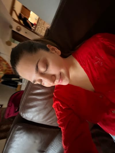 The scene is located in a residence in Ancenis, France. A young individual with dark hair, pulled back from the face, is depicted lying on their left side on a brown leather sofa. Their eyes are closed, and a slight smile is visible. They are wearing a long-sleeved red top with a textured or patterned fabric. In the blurred background, another person, dressed in a dark top, is partially visible within an adjacent, brightly lit indoor area. This background space suggests a domestic setting, possibly a kitchen or dining area, with light-colored walls and a tiled surface visible.