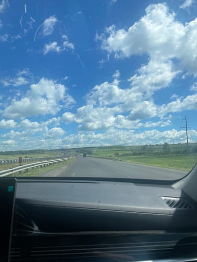 This image captures a view from inside a vehicle traveling on a highway. The sky is a vibrant blue, dotted with fluffy white clouds. The highway stretches ahead, flanked by grassy fields and trees on either side. Power lines with tall pylons are visible in the distance. The dashboard of the car is partially visible in the foreground, suggesting the perspective of a driver or passenger. The image appears to have been taken on a sunny day. The provided context "city of Kazyli, Russia" does not appear to be verifiable from the image content alone.