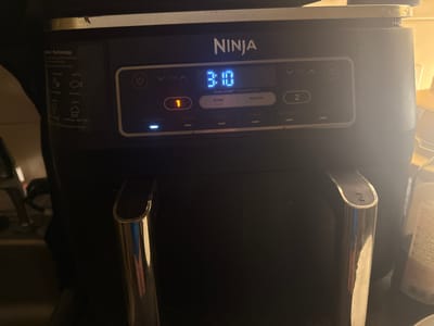 The image shows a close-up of a black Ninja DualZone Air Fryer on a countertop in Cheltenham, United Kingdom. The air fryer has a digital display showing "3:10" and illuminated buttons for "MAX CRISP", "AIR FRY", "ROAST", "REHEAT", and "DEHYDRATE". There are also buttons for "SYNC" and "MATCH", and two numbered zones, "1" and "2". The appliance is positioned next to a sink and a bottle of liquid. The lighting suggests an indoor environment, possibly a kitchen.