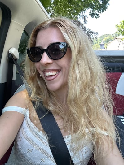 The image shows a blonde woman with sunglasses and a white lace top inside a car. She is smiling and looking to her left. A black seatbelt is visible across her chest. Outside the car, trees and a building with a steeple can be seen, suggesting a rural or suburban setting. The location is identified as Schoenfels, Luxembourg.