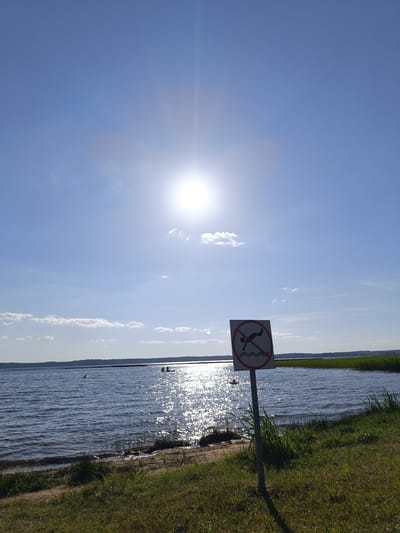 This media file depicts a scenic view of a body of water, likely a lake, on a bright, sunny day. The sun is high in the sky, casting a brilliant glare on the water's surface, creating a shimmering effect. A few scattered clouds dot the otherwise clear blue sky.
In the foreground, the shoreline is lined with green grass and some patches of sand. A prominent sign is posted on a pole, featuring a red circle with a diagonal line through it, indicating a prohibition. Inside the circle, a pictogram depicts a person diving into water, signifying that diving is not allowed in this area.
Further out on the water, several individuals are visible. Some appear to be swimming or wading, while others are in small boats or on rafts, enjoying the water. The overall atmosphere suggests a recreational setting, likely a popular spot for outdoor activities.
The presence of the "no diving" sign suggests caution is advised in this location, possibly due to unknown water depths or submerged hazards. The time of day is clearly midday or early afternoon, given the sun's position and the bright illumination. The lush green vegetation on the shore hints at a temperate climate, and the location context provided points to Belarus. The overall mood is peaceful and inviting, despite the warning sign.