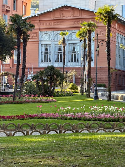 The image shows a park in Opatija, Croatia. The park is next to a salmon-colored building. Palm trees line the park's edge, and there are rows of pink and white flowers alongside a decorative stone fence. The grass is well-maintained and green. People can be seen walking in the distance. Street lamps are visible among the palm trees. The building has many windows and is made of brick or a similar material.