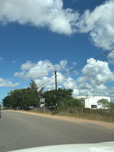 The image shows a road in what appears to be a rural area, possibly near or in the city of Muxara, Mozambique. A gray asphalt road stretches into the distance, bordered by green vegetation and a few scattered trees. On the left side of the road, utility poles and power lines are visible, extending upwards toward a sky filled with scattered white clouds against a blue backdrop. A white building is also visible in the background. The photograph appears to have been taken from inside a vehicle, with part of the car's interior visible on the right side of the frame. There are no people or other subjects immediately apparent in the scene. The overall environment conveys a sense of a quiet, possibly sparsely populated area.