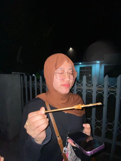 A young woman wearing a brown hijab and glasses is standing outdoors at night, holding a skewer with food in her right hand and a smartphone in her left. She appears to be taking a selfie with her phone. The background includes a white fence, a building with lights, and dark foliage, suggesting a residential area in Tanggul, Indonesia. The lighting is dim, with artificial lights from the building and possibly streetlights.