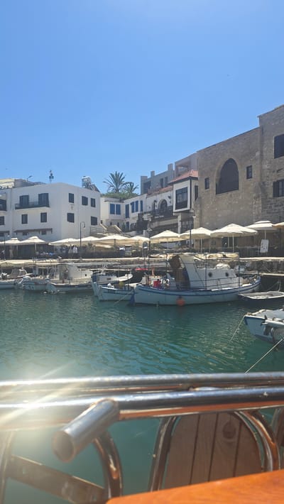 The image shows a bright, sunny day in the Kyrenia harbor in Northern Cyprus. Several small fishing boats are docked in the clear blue water. In the background, white buildings with blue shutters line the harbor, and a large stone building with an arched entrance stands to the right. Outdoor seating areas with white umbrellas are set up in front of the buildings. A wooden railing and part of a wooden chair are visible in the foreground, suggesting the photo was taken from a boat or a similar vantage point. The water is slightly rippled, and sunlight reflects off its surface. A palm tree is visible behind some of the white buildings. The sky is a clear, vibrant blue.