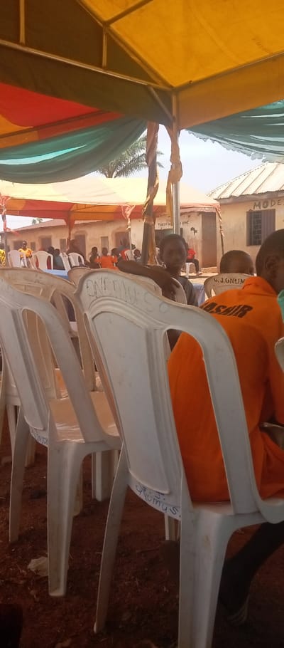 The image captures an outdoor gathering in Kasuwan Magani, Nigeria, under a multi-colored canopy. The canopy features panels of yellow, red/orange, and green/teal fabric supported by a visible metal framework, with some support poles wrapped in brown material. Rows of white plastic chairs are arranged on a reddish-brown earth ground.
In the right foreground, the back of a person wearing an orange shirt with "BASHIR" printed in black capital letters is visible, seated in a white plastic chair. To the left, a young individual with dark skin, wearing a dark t-shirt, is seated in a white plastic chair, looking directly at the camera. Their left arm rests on the back of an adjacent chair. Visible inscriptions on the white plastic chairs include "B.J. ORUM" on a chair in the midground and "J. S. QROiuS." on a chair to the right.
Further into the background, beneath additional sections of the canopy, numerous other individuals, including children, are seated in similar white plastic chairs. Beyond the canopy structure, several single-story buildings with light-colored walls and corrugated metal roofs are visible. One building in the mid-right background has "MODE" (likely part of "MODEL") painted in black letters on its wall, while another building in the mid-left background shows partial text "TUREY H...". A palm tree is visible against a bright, overcast sky in the distance. The ground beneath the seating area is exposed reddish-brown soil, consistent with laterite. The scene suggests a community event or assembly.