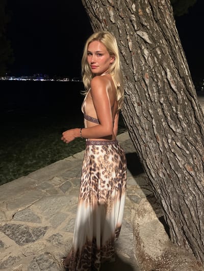 A blonde woman in a leopard print dress stands next to a large tree in Port de Pollença, Spain. The woman is facing away from the camera, looking over her shoulder. She has a bracelet on her left wrist. The dress is long and flowing, with a high slit. The tree is rough-barked and appears to be an olive tree. In the background, the lights of the town can be seen across the water. The scene is lit by artificial lights, suggesting it is nighttime. The ground is paved with stone.
