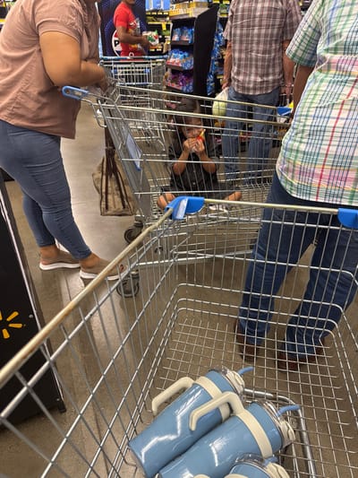 The image depicts an interior scene at a Walmart supermarket in La Ceiba, Honduras. In the immediate foreground, a metal shopping cart contains three light blue insulated tumblers, each with a light tan handle, a clear lid, and a blue straw. Directly behind this, a second metal shopping cart holds a young child, appearing to be female, with dark hair, sitting cross-legged on the cart's base. The child is wearing a dark shirt and is seen holding a small, red, fruit-like object to her mouth, actively eating it.
To the left, a woman is partially visible, dressed in a light brown, short-sleeved top and blue jeans, wearing brown slip-on shoes with white soles. She is positioned next to a shopping cart, with a brown handbag hanging nearby. Further to the right, an individual is partially visible from the waist down, wearing a brown plaid shirt, blue jeans, and brown shoes, standing behind another shopping cart. On the far right, a person's lower torso and legs are visible, wearing a green, white, and red plaid shirt and blue jeans, also near a shopping cart. In the background, retail shelves stocked with various packaged goods are visible, and another individual in a red shirt is seen holding a plastic bag near a distant shopping cart. The floor is grey concrete, and a partial yellow starburst logo, consistent with Walmart branding, is visible on a black display stand on the far left.