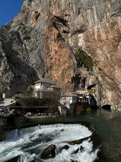 The image depicts a panoramic view of the Blagaj Tekke complex situated at the base of a large, craggy limestone cliff face in Dračevice, Bosnia and Herzegovina. The cliff, which dominates the background, exhibits varied grey, brown, and reddish-orange geological strata, with sporadic green vegetation in crevices, and a large cave entrance visible at its base on the far right.
In the mid-ground, a cluster of traditional Ottoman-era buildings with white walls, stone roofs, and distinctive wooden-framed windows, including an overhanging bay window, are constructed into and alongside the cliff. To the left of these main structures, at river level, an outdoor restaurant area with a dark-colored canopy covers multiple tables and chairs. Several indistinct individuals are seated at these tables.
In the foreground, the emerald-green Buna River flows from right to left, emerging from the cave at the base of the cliff. A man-made weir or dam spans the river, creating a wide waterfall of turbulent, white-foamed water and rapids over dark rocks in the immediate foreground. The water upstream of the weir is calmer. Bare deciduous tree branches are visible near the river's edge, suggesting a late autumn or winter season. The sky in the upper left is clear blue, indicating fair weather conditions.