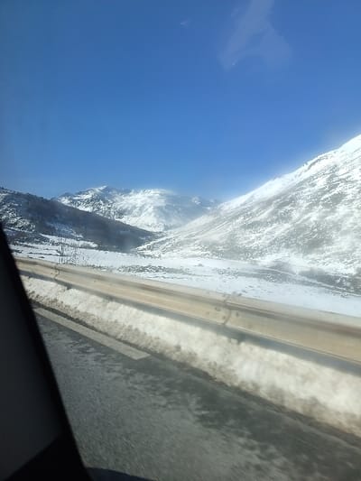 A snowy mountain landscape stretches under a clear blue sky. The road, visible with a roadside barrier, is damp and reflects the light. Snow is piled up along the side of the road. The mountains are covered in a thick blanket of snow, with some bare trees and rocks visible. The perspective indicates the image was taken from inside a moving vehicle.
