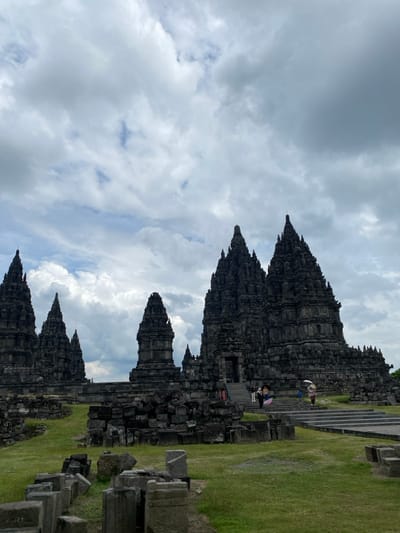 The image shows the Prambanan Temple complex, located in the city of Kecamatan Prambanan, Indonesia. The temple complex consists of several large stone structures, with the main temple in the center. The structures are made of gray stone and are adorned with intricate carvings. The image is taken from a distance, looking towards the front of the temple, with a large area of grassy lawn in the foreground, and several smaller stone structures scattered across the lawn. The sky is overcast with clouds, and a few people can be seen walking on the steps leading up to the main temple. The scene is serene and peaceful, with the ancient temple complex standing as a testament to the rich history and culture of Indonesia.