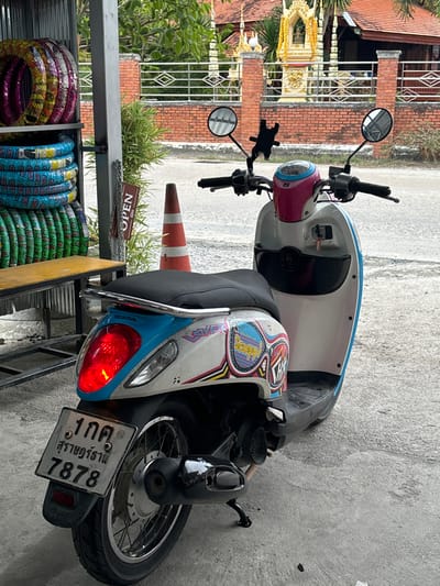 A white, pink, and light blue Honda Scoopy scooter is parked on a concrete surface, angled slightly to the right, with its kickstand deployed. The scooter features a black seat, a silver rear grab rail, black mirrors, and a black smartphone holder mounted on its handlebars. Its red taillight is illuminated. Colorful, cartoonish decals, including "Scoopy!" text, adorn the rear side panels. The Thai license plate at the rear reads "1 กค สุราษฎร์ธานี 7878".

To the left of the scooter, a metal display rack holds stacks of new, brightly patterned tires in pink/yellow, blue, and green. Below these tires is a bench with a yellow top. A brown "OPEN" sign, written in both Thai and English, hangs from a metal support post. An orange and white traffic cone stands near the sign.

In the background, beyond an asphalt road, a red brick fence encloses a property. Prominently centered behind the fence is a golden, ornate spirit house adorned with a yellow garland. Green foliage, including bamboo and other trees, is visible around the fence and in the distant background. No individuals are present in the scene. The image depicts an outdoor roadside setting in Baan Nai Wok, Thailand.
