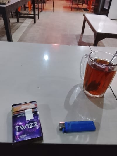 The image depicts a static scene within an establishment located in Dayahlhoksukon Barat, Indonesia. In the immediate foreground, a light-colored table surface occupies the lower half of the frame. On this table, a purple pack of "TWIZZ ROYAL CROWN" cigarettes is positioned on the left, with a visible "PERINGATAN" (warning) label near the top. To its right, a blue disposable lighter rests horizontally. Further to the right, a clear glass mug containing a dark brown liquid, presumably tea, holds a metal spoon.
The midground reveals a light-toned floor with a distinct white line traversing it horizontally. Several dark-legged tables with light-colored tops are visible in this area, suggesting a dining or seating arrangement. One such table is partially visible on the far left, while another with a white top and dark legs is prominent in the center-right, set back from the foreground table. The background features blurry, warm-toned lighting, indicating artificial illumination or ambient light, with indistinct shapes of further furniture or structures. No individuals are present in the frame.