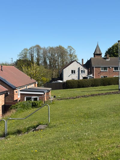 This media file captures a sunny, clear day in a residential area of Bearpark, United Kingdom. The scene features a row of houses bordered by a wide, green grassy area.
On the left side of the image, several houses are visible. One prominent house in the mid-ground features red brick walls and a distinctive dark, pointed, or conical roof structure, resembling a small spire or turret. Below this, another house has light-colored walls and a dark roof. In the foreground, partially visible, is a house with a reddish-brown tiled roof and brickwork, displaying a television aerial. Between the houses and the grassy expanse runs a neatly trimmed green hedge.
The right side of the image is dominated by a large, well-maintained lawn, dotted with small white and yellow wildflowers, likely daisies and dandelions. A metal railing, possibly indicating a path or boundary, is present in the lower right foreground next to the grass. In the background, numerous deciduous trees, mostly bare or with very early foliage, rise against a bright, cloudless blue sky, which fills the upper portion of the image. A street light is visible near the top right.
The overall impression is one of a quiet and peaceful suburban or village setting under bright daylight. No human activity or visible text is present.